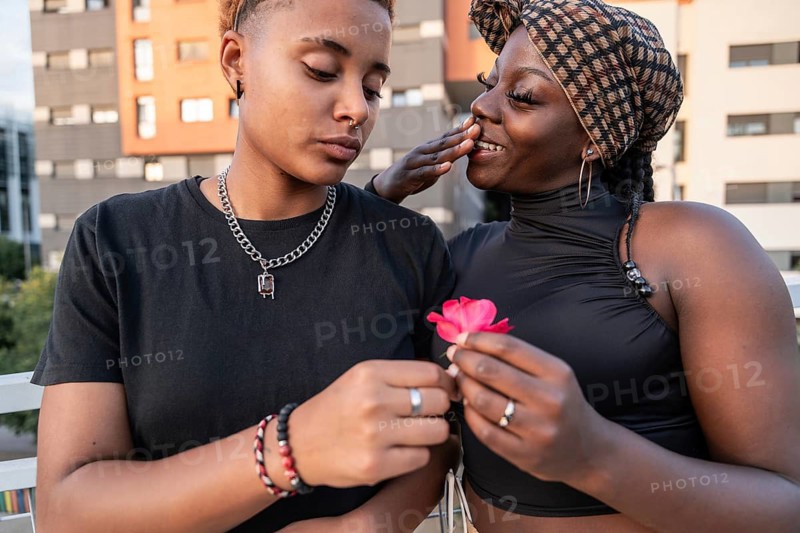 A joyful LGBT multiethnic couple shares a tender moment, smiling warmly at each other while holding a vibrant pink flower against an urban backdrop