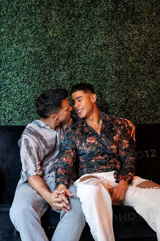 A loving latin gay couple sits closely on a sofa, holding hands and sharing a joyful, intimate moment with smiles, against a lush green plant wall, expressing warmth and affection