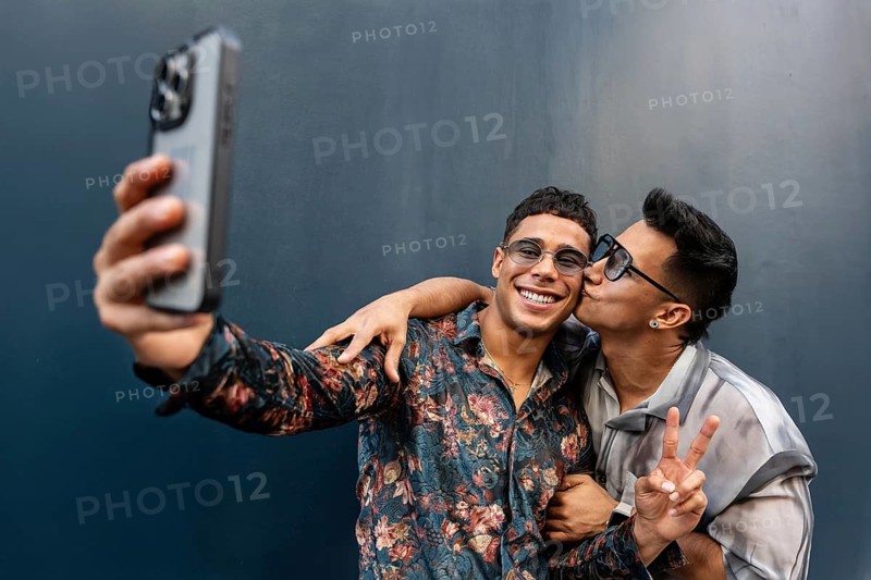 A joyful latin gay couple takes a selfie, capturing a moment of affection and happiness. One person kisses the other on the cheek, while making a peace sign