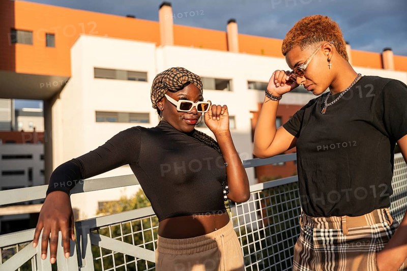 A fashionable LGBT multiethnic couple poses confidently in the sunshine, leaning against a railing. The modern architecture in the background adds to the vibrant urban scene