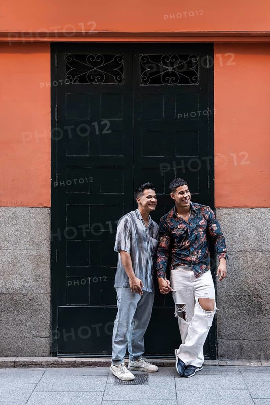 A joyful latin gay couple holds hands while standing in front of a colorful building in Madrid. The scene captures love, diversity, and cultural vibrancy