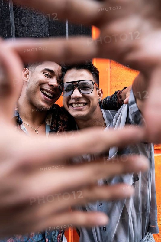 A joyful latin gay couple poses with smiles, framing their faces with their hands for a selfie against a vibrant orange wall, capturing a moment of happiness and love