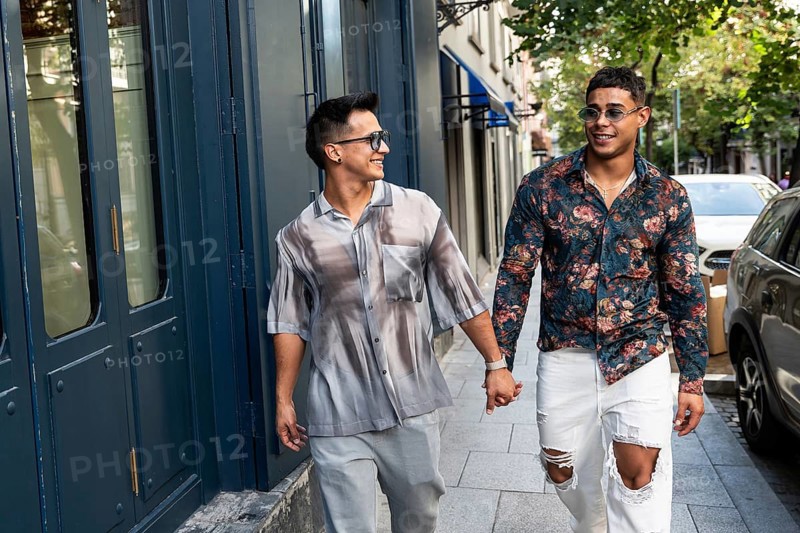 A happy latin gay couple holds hands while strolling down a lively city street in Madrid. They wear stylish outfits and smile warmly at each other, enjoying a sunny day