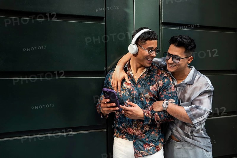 A joyful latin gay couple embraces while listening to music. One wears headphones and a floral shirt, the other sports a watch and sunglasses, expressing love and happiness