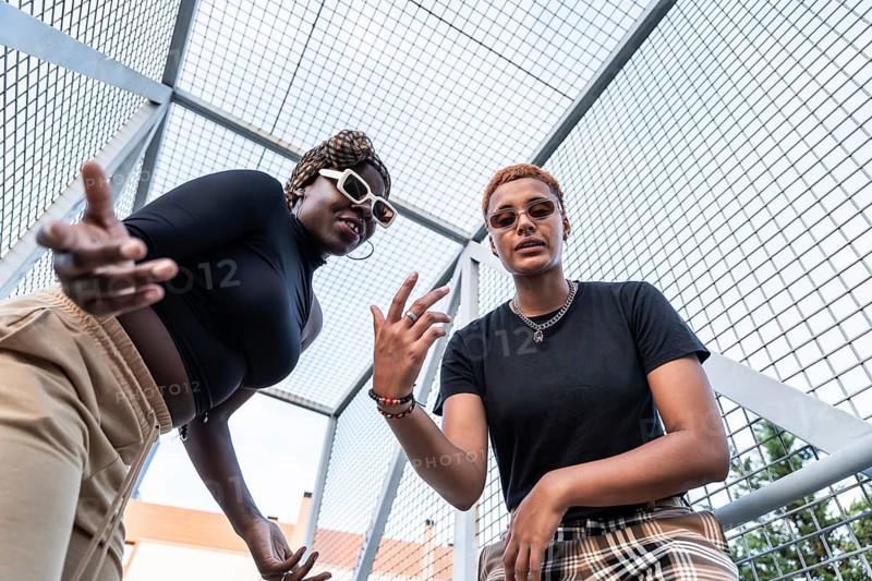 A dynamic LGBT multiethnic couple poses confidently in an urban setting. Both showcase unique styles with sunglasses and casual outfits, highlighting individuality and modern expression