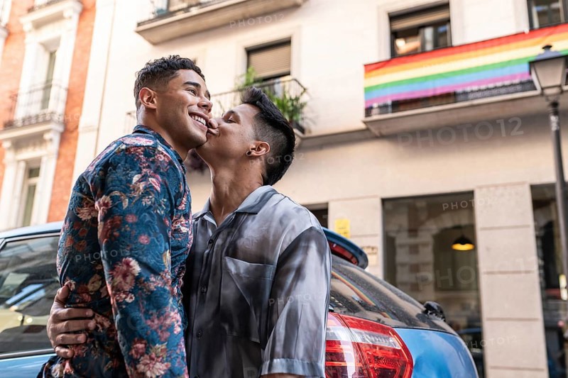 A joyful latin gay couple shares a tender moment on a lively street in Madrid, with a rainbow flag in the background, symbolizing pride and love