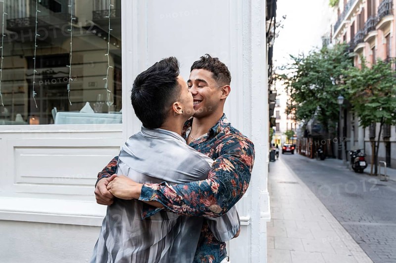A joyful latin gay couple embraces and shares a tender moment on a charming street in Madrid. The vibrant city backdrop adds to the warmth of their connection and happiness