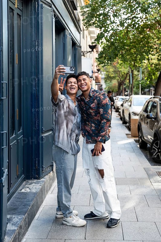 A happy latin gay couple taking a selfie on a bustling street in Madrid. They smile and embrace, capturing the vibrant atmosphere and their joyful connection