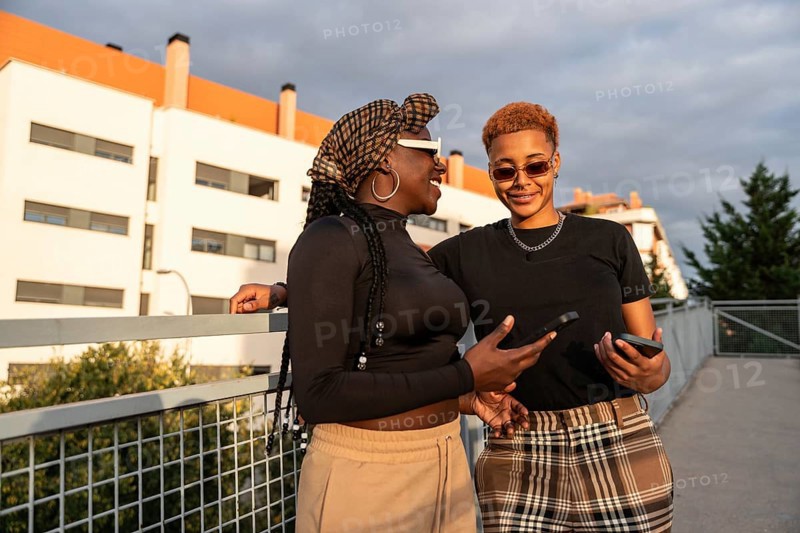 A happy LGBT multiethnic couple smiling while holding phones, enjoying the sunny outdoors in fashionable casual clothing, standing on a bridge with modern buildings in the background