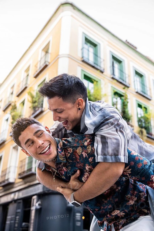A joyful latin gay couple share a playful piggyback ride in the vibrant streets of Madrid. The warm atmosphere captures their happiness and connection, set against a charming urban backdrop