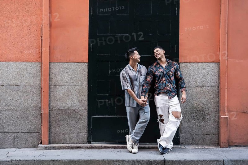 A joyful latin gay couple shares a tender moment holding hands, standing in front of a colorful urban backdrop in Madrid, Spain, capturing the essence of love and happiness