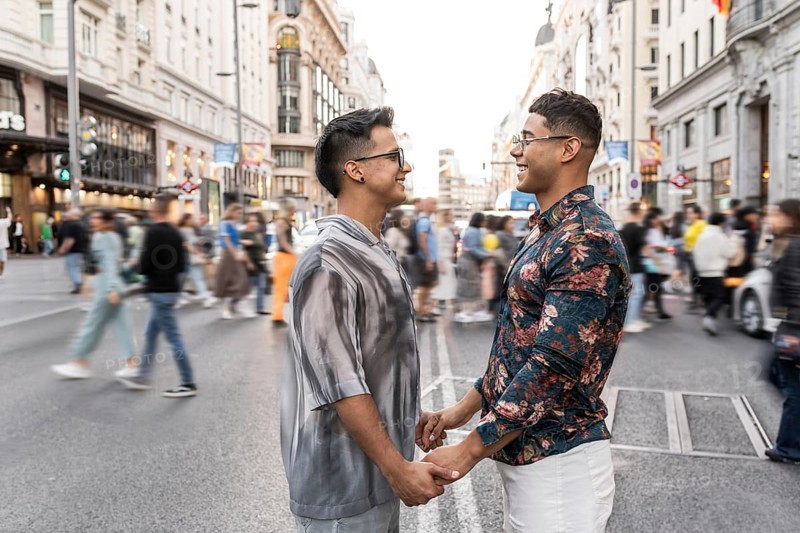A joyful latin gay couple holds hands and smiles at each other amidst the hustle and bustle of a busy street in Madrid, depicting love and connection in an urban setting