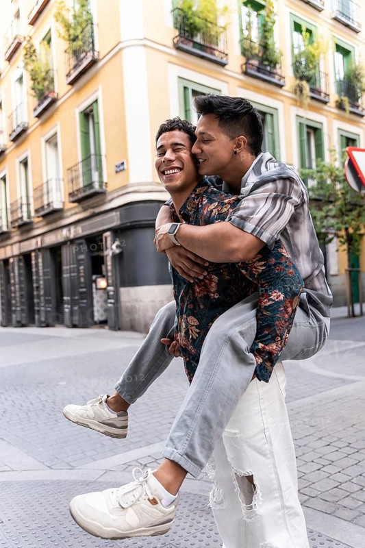 A joyful latin gay couple piggybacking on a picturesque street in Madrid. The colorful buildings and cobblestones add charm to their happiness