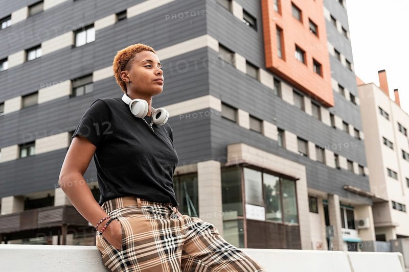 A confident young hispanic transgender with short hair wearing headphones and plaid pants, leaning casually against a railing in an urban setting