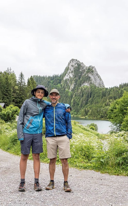 A father and son enjoy a hiking adventure at Lac de Taney, Switzerland, surrounded by lush greenery and a majestic mountain backdrop The scene captures the joy of nature exploration
