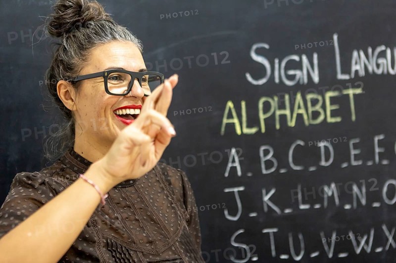 A woman teaches sign language alphabet in a classroom, gesturing with a smile Chalkboard filled with letters in the background adds an educational atmosphere