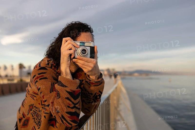 Casual moment of a young man enjoying photography by the scenic waterfront He is dressed in a cozy patterned sweater, holding a camera, against a serene backdrop