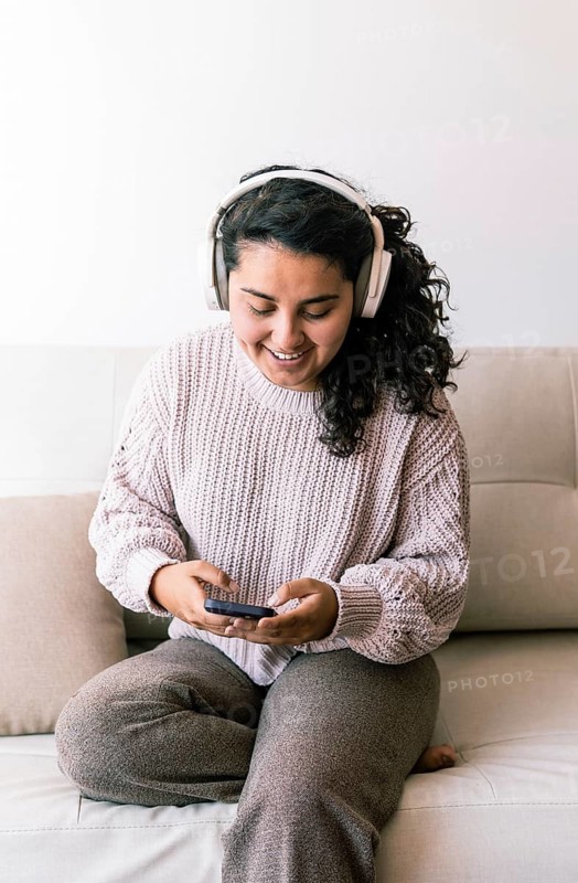 A young latin woman sits comfortably on a couch, wearing headphones and looking at her phone She exudes relaxation and happiness, enjoying her time at home