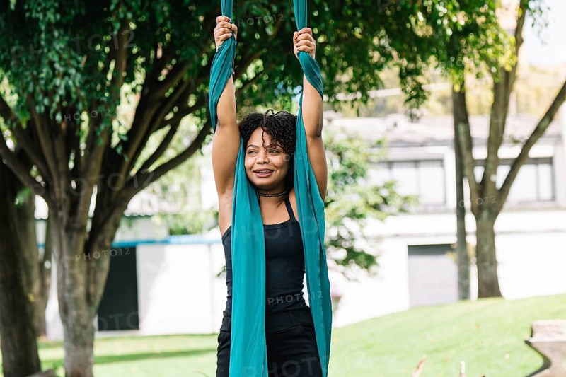 A black performer practices aerial gymnastics on fabric in a vibrant Bogota park, surrounded by lush trees The scene captures the grace and strength of this art form