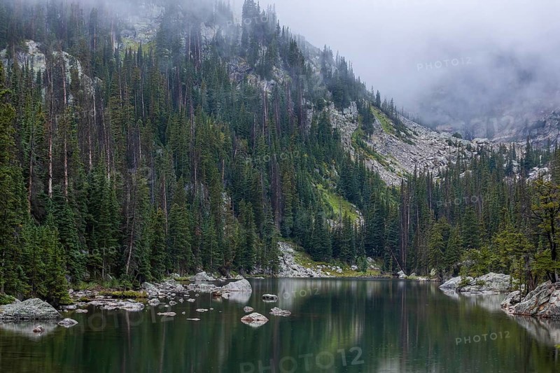 A tranquil mountain lake in Colorado, shrouded in mist, nestled within dense, towering pine forests and rugged terrain, highlighting the serene beauty of nature in the Rocky Mountains