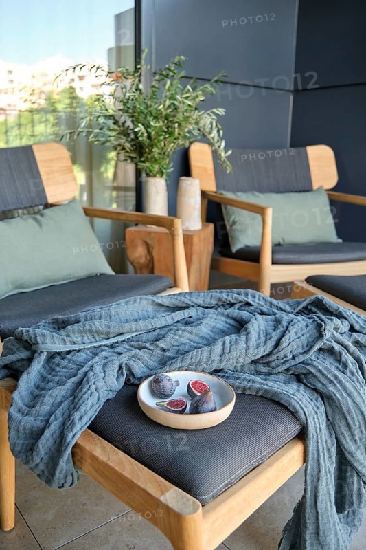 A stylish modern balcony featuring wooden chairs with cushions, a knitted throw, and a small table with a bowl of figs, complemented by a potted plant and neutral background