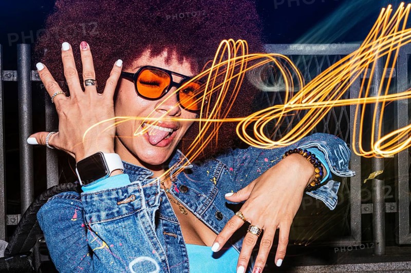 African descendant woman with afro hair showcases her playful side, winking and sticking her tongue out with orange-tinted sunglasses at night. Light trails swirl in the background, adding a dynamic and whimsical atmosphere