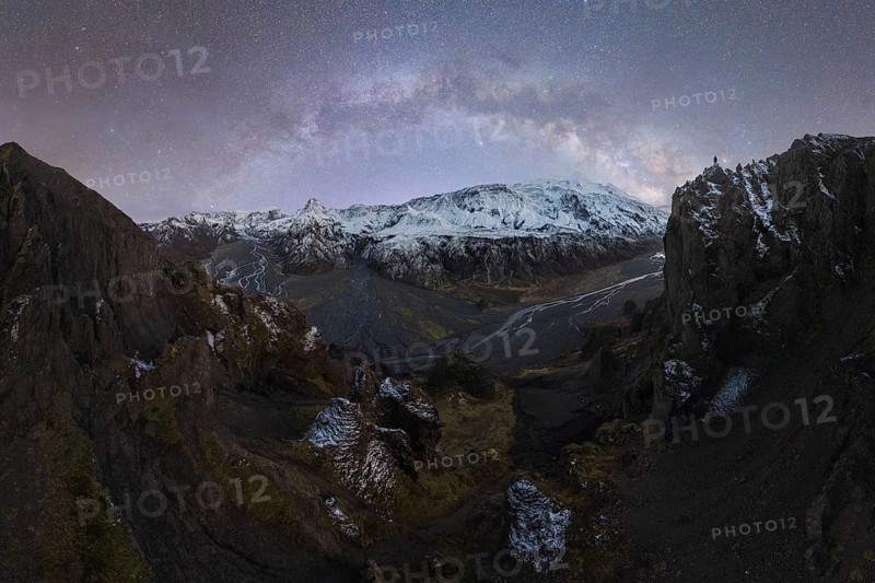 View of Icelandic peaks under a starry night, with the Milky Way arching over the snowy landscape Rugged terrain and river patterns add to the scene's beauty A lone figure stands on one of the peaks