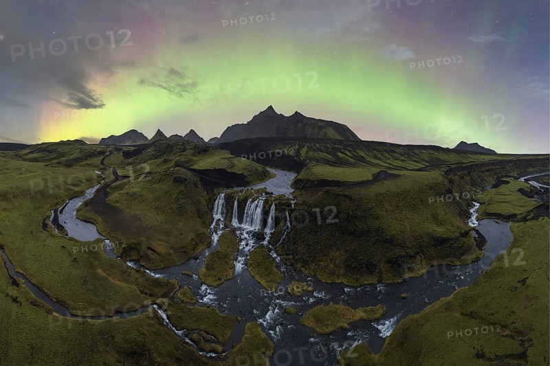The aurora borealis illuminates the sky above a stunning Icelandic waterfall and meandering river surrounded by rugged terrain
