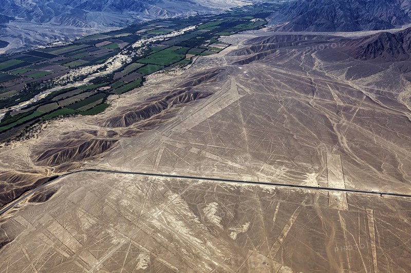 Aerial photo shows dry geoglyphic landscape next to green fields and a road, the geoglyphs and drawings in the desert near Nasca and Palpa in Peru