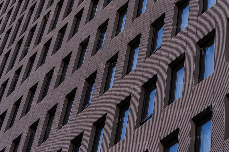 A detailed view of a modern building facade featuring a repetitive geometric pattern with aligned windows set in dark, textured cladding. This architectural design emphasizes modern aesthetics and urban style