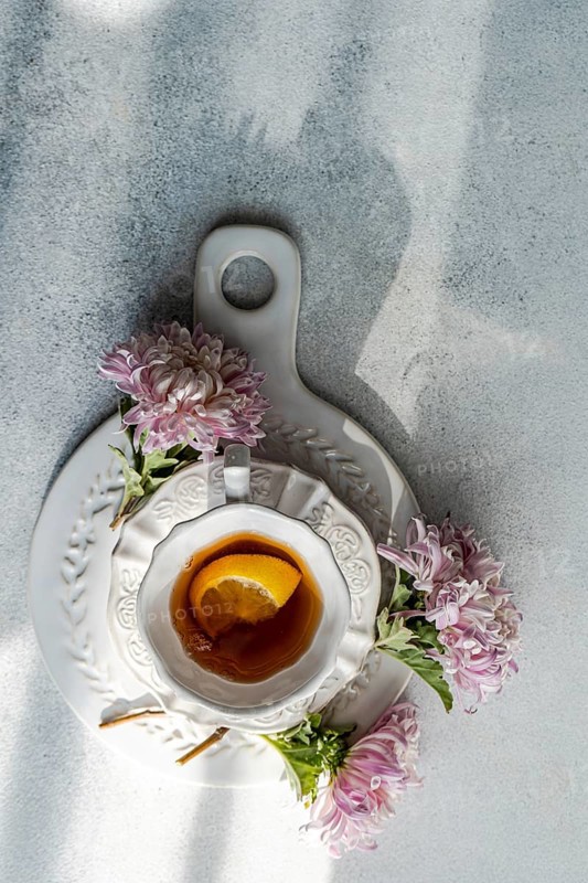Aerial view of a rustic autumn tea setup featuring a white cup and saucer on stone, garnished with lemon slice and flanked by delicate pink chrysanthemums