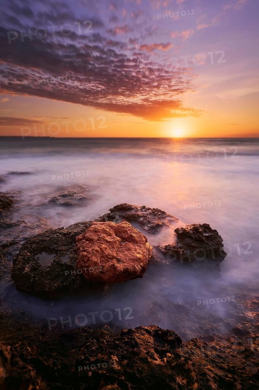 A breathtaking sunset scene at Cabo Roig, Orihuela Costa, Alicante, with vibrant colors reflecting on calm waters and textured rocks in the foreground, creating a tranquil ambiance