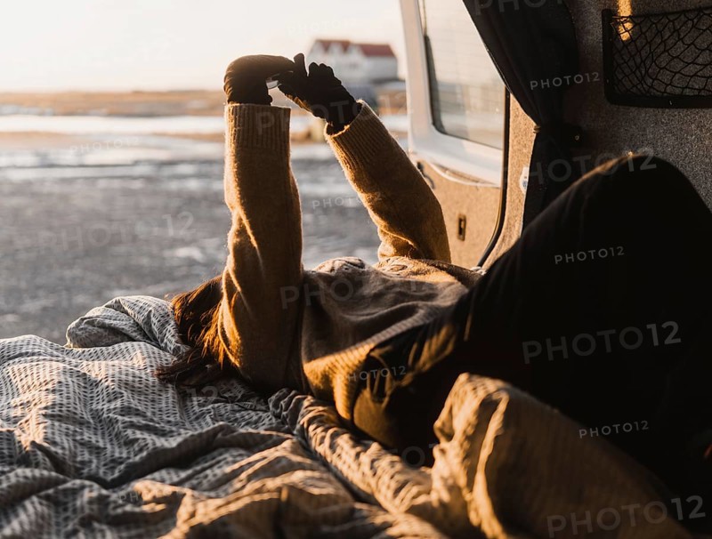 Unrecognizable woman lounging in a van during a camping trip in Iceland. She stretches her arms against the sunlight, creating a cozy, warm atmosphere