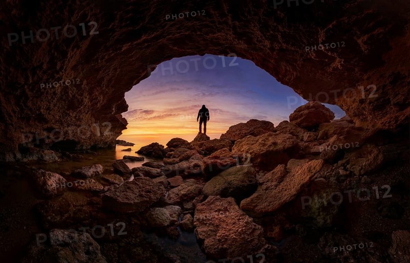 A stunning silhouette of a person standing on rocky terrain under a vibrant sunset sky, viewed from a cave at Minas Romanas, Torre de la Horadada, Alicante, Spain