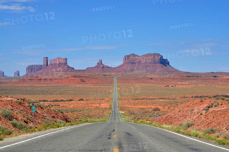 The long, straight road runs through a desert-like landscape with rocks. Access to Monument Valley, Navajo Tribal Park, Navajo Nation Reservation, Utah, Arizona, USA