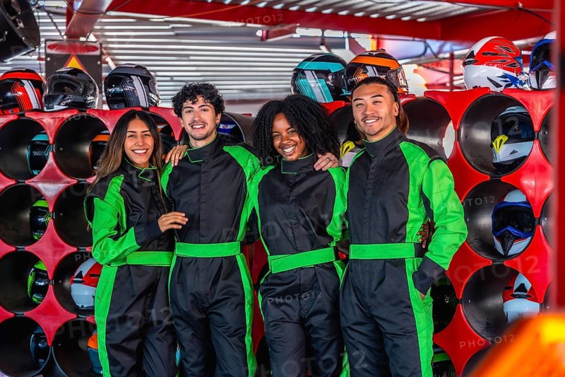 Happy diverse friends smiling, wearing green racing suits, standing together in a karting facility with helmets stored in the background, sharing a fun and exciting racing experience