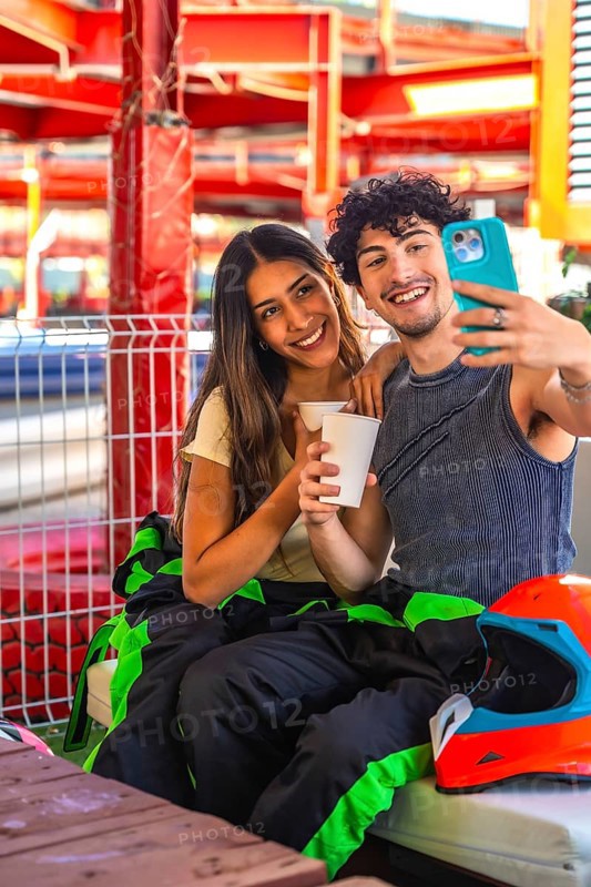 Young couple wearing racing suits taking a selfie during a karting break, showing happiness and connection as they enjoy a recreational activity together