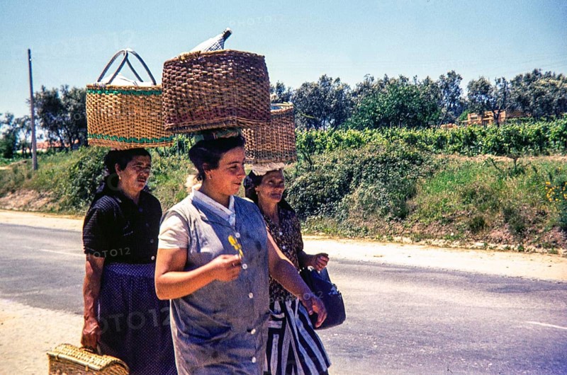 Three women walking on street in rural area carrying wicker baskets on their head, Portugal, Europe 1968