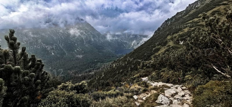 Wild mountain trail with a view of a lake, Morskie Oko and thick, dramatic clouds above the peaks, hiking in the High Tatras National Park, Carpathian Mountains, Poland