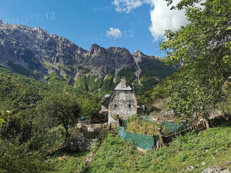 Stone house surrounded by gardens and mountains under a clear blue sky, Peaks of the Balkans, hiking in the Albanian Alps National Park, Albania
