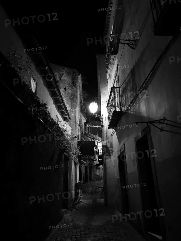 A narrow alley at night with illuminated alleyway lamp between tall, dark buildings, monochrome, atmospheric, Lisbon, Portugal