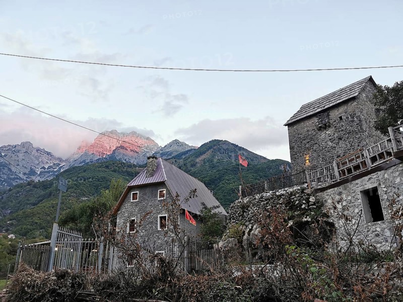 Traditional village with stone houses and Albanian flags against a mountain backdrop, mountain glow, alpine glow in the background, Peaks of the Balkans, hiking in the Albanian Alps National Park, Albania