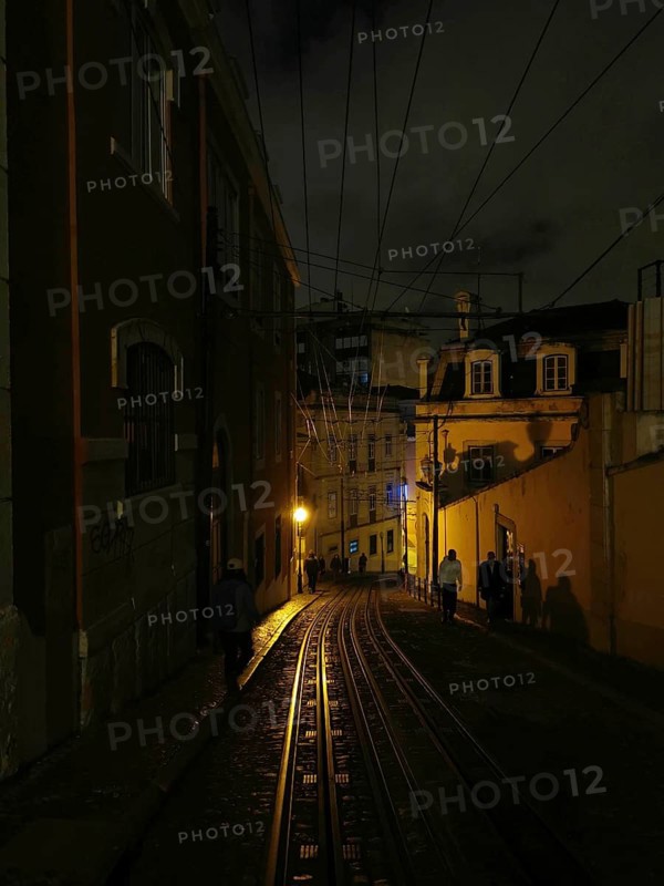 Rails of the Elevador da Gloria funicular, A dark alley at night with lantern light and shadows from passers-by, Lisbon, Portugal