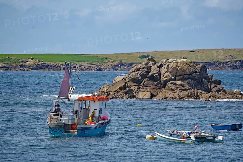 A small fishing boat out at sea off a rocky coast with colorful sails and clear skies, St Agnes, Isles of Scilly, Cornwall, Great Britain