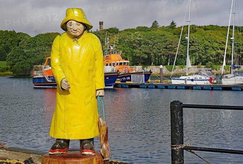 Statue of a fisherman in yellow raincoat at harbour in front of anchored boats with green hills in the background, Stornoway, Hebrides, Scotland, Great Britain