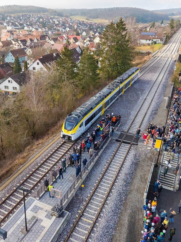 A train stops at a platform surrounded by a crowd of people. In the background you can see a village and wooded hills, opening of the Hermann Hesse Railway, Calw, Germany