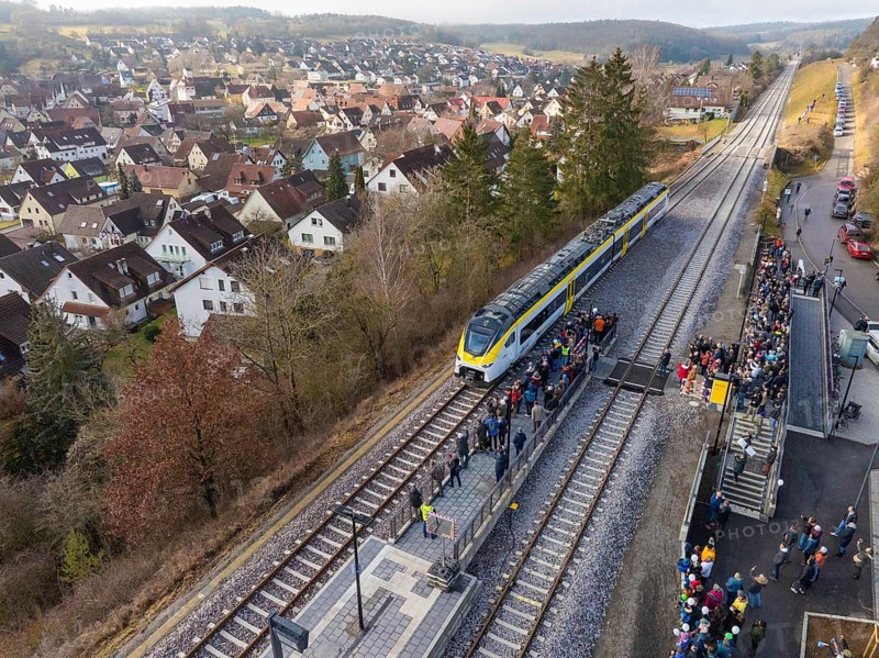 A train stops at a rural train station with surrounding people. The surrounding area shows a village and hills, opening of the Hermann Hesse Railway, Calw, Germany