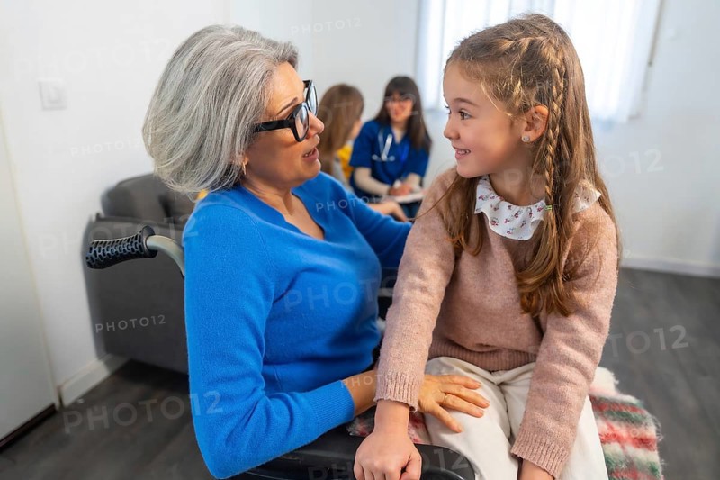 Grandma in a wheelchair interacting happily with her granddaughter, sharing a warm moment during a family visit at a facility with healthcare professionals present in the background
