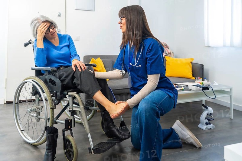Medical professional providing physical therapy and attentive care to an elderly patient experiencing discomfort in her leg while sitting in a wheelchair at home