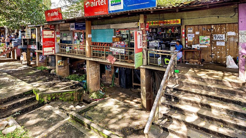 A storefront with various signs and posters surrounded by trees and a rustic atmosphere, The old market halls of Kandy in Sri Lanka